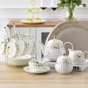 Op de afbeelding: Een witte porseleinen theeset met een bloemenpatroon. De set bevat een theepot, suikerpot, melkkan, theekopjes, schotels en lepels. De kopjes hangen aan een goudkleurig metalen rek.