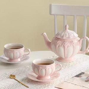 Puede incluir: Un juego de té de cerámica rosa y blanca, que incluye una tetera y dos tazas con platillos. El juego presenta un diseño en relieve detallado. Las tazas y la tetera están llenas de té. Una cuchara dorada descansa sobre la mesa.
