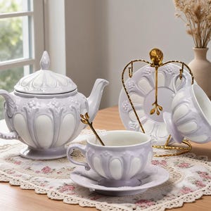 Könnte beinhalten: Ein Keramik-Teeservice bestehend aus einer Teekanne, zwei Tassen und zwei Untertassen in Helllila und Weiß mit geprägten Blumendetails. Das Set enthält zwei goldfarbene Löffel und einen goldfarbenen Metallständer für die Tasse und Untertasse.