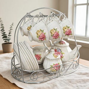 Puede incluir: Un juego de té que incluye tazas, platillos, una jarra de leche y una tetera de porcelana blanca con motivos florales de rosas rosas y amarillas y bordes dorados, organizados en un soporte de alambre metálico plateado con asa.