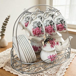 Può includere: Un supporto in filo metallico argentato che sostiene un servizio da tè in porcellana bianca. Il set comprende tazze, piattini, una lattiera, una zuccheriera con coperchio e una teiera, decorati con motivi di rose rosa e piccole farfalle.