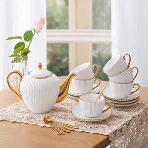 Puede incluir: Un juego de té de cerámica blanca que incluye una tetera, cuatro tazas con sus platos y cuatro cucharas doradas. Cada pieza presenta una textura acanalada vertical y bordes, asas y boquillas con detalles en color dorado sobre un tapete.