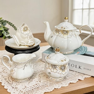Peut inclure: Un service à thé en porcelaine blanche comprenant une théière, un pot à lait, un sucrier avec couvercle et une tasse sur soucoupe. Chaque pièce est ornée de motifs floraux dorés. La théière présente un bec verseur et un couvercle doré.