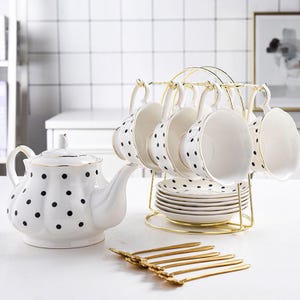 Puede incluir: Un juego de té de porcelana blanca con lunares negros, que incluye una tetera, tazas, platillos y un soporte de metal dorado. El juego está dispuesto sobre una superficie blanca, con cucharas doradas en primer plano.