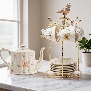 Peut inclure: Une théière en porcelaine blanche et un ensemble assorti de tasses et soucoupes ornées de motifs de papillons et de fleurs colorés avec des bordures dorées. Les tasses sont suspendues à un support en métal doré surmonté d'un petit oiseau.