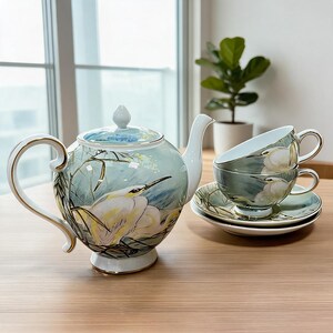 May include: A porcelain teapot and two matching teacups with saucers, featuring a light blue and green background with painted white egret birds and reeds. Each piece is finished with a delicate gold trim along the edges and handles.
