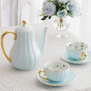 Puede incluir: Juego de té de porcelana azul claro con detalles dorados. El juego incluye una tetera, dos tazas y platillos. Las tazas y los platillos tienen un diseño floral. Un jarrón con flores azules se encuentra en el fondo.