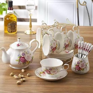 Op de afbeelding: Een witte porseleinen theeservies met rozenmotieven. De set bevat een theepot, kopjes, schotels en een goudkleurig metalen rek. De kopjes en schotels zijn op een houten tafel geplaatst.