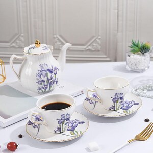 Op de afbeelding: Een wit porseleinen theeservies met een theepot, twee kopjes en schotels. De set is versierd met paarse iris bloemmotieven en gouden randen. Eén kopje is gevuld met donkere vloeistof. De set staat op een wit oppervlak.