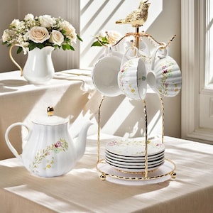 Op de afbeelding: Een witte porseleinen theeset met bloemmotieven. De set bevat een theepot, kopjes, schotels en een decoratieve standaard met een gouden vogel. Een witte kan met bloemen is ook aanwezig.