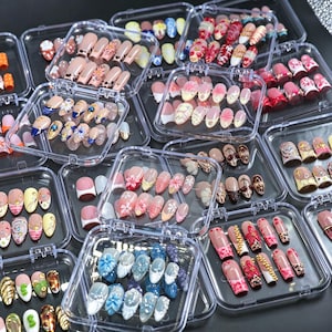 May include: A collection of multiple clear plastic cases containing various sets of press-on artificial nails. The nails feature diverse designs including floral patterns, leopard prints, metallic accents, rhinestones, and solid colors in various shapes and lengths.