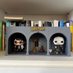 May include: A gray, brick-style display shelf featuring Harry Potter characters. The shelf has three arched openings, each housing a figure. The Harry Potter logo is centered above the arches. Books and other items are on the shelf above.