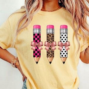 May include: A pale yellow t-shirt featuring a graphic of three pencils arranged side-by-side. Each pencil has a pink eraser, a light pink bow tied around the center, and a unique pattern: one with a black and pink checkerboard, one with a brown leopard print, and one with black polka dots.