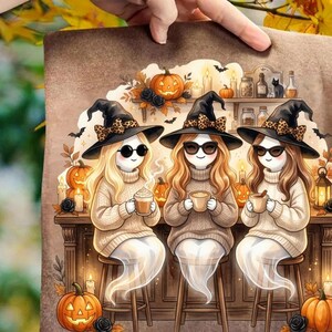 May include: A graphic print featuring three ghost figures wearing black witch hats with leopard print bows, sunglasses, and beige cable knit sweaters. They sit on stools holding mugs. The background shows shelves with pumpkins, candles, and jars.