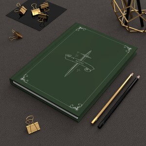 May include: A dark green notebook with a white sword and banner design that reads "NOTES". The notebook has a decorative border and is accompanied by gold and black pencils and gold binder clips. The notebook is on a dark gray surface.