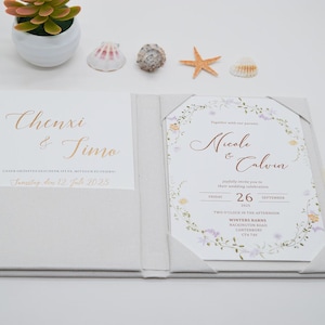 May include: A wedding invitation suite with a floral design. The invitation features the names "Nicole & Calvin" and the date "26 September 2025". The suite includes a pocket-style holder with the names "Chenxi & Timo" printed in gold.