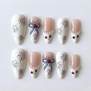 May include: A set of ten artificial nails with a white base, featuring red, white, and blue firework designs. Some nails have a nude pink base with pearl and star accents, and a bow design with red and blue stripes.