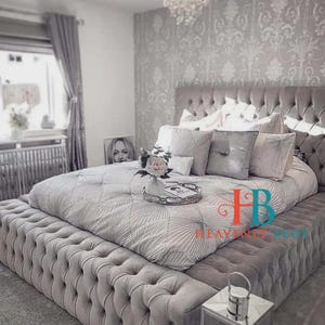 May include: A luxurious, tufted bed in a bedroom setting. The bed frame and headboard are upholstered in a soft gray fabric. The bedding includes a patterned comforter and several decorative pillows. A small tray with flowers sits on the bed.