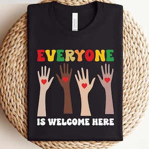 以下が含まれることがあります： 「EVERYONE IS WELCOME HERE」というカラフルで丸みを帯びたフォントのテキストがプリントされた黒いTシャツ。テキストの下には、さまざまな肌の色の4つの手が上げられており、それぞれの掌の中央に小さな赤いハートが描かれています。