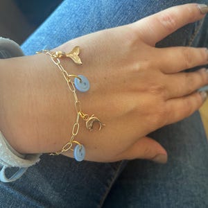 Könnte beinhalten: Ein goldfarbenes Gliederkettenarmband mit zwei hellblauen runden Anhängern und zwei goldfarbenen Anhängern, einer in Form eines Delfins und der andere in Form einer Meerjungfrauenflosse, die mit kleinen goldfarbenen Ringen befestigt sind.