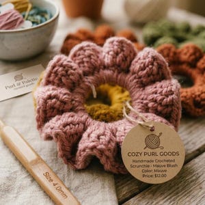 Könnte beinhalten: Ein handgefertigtes, gehäkeltes Haargummi in Mauve mit einem gelben Zentrum, das einer Blume ähnelt. Das Haargummi wird mit einer Häkelnadel und einem Etikett mit der Aufschrift "COZY PURL GOODS" gezeigt. Weitere Haargummis in verschiedenen Farben sind im Hintergrund zu sehen.