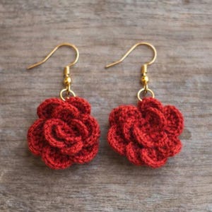 Könnte beinhalten: Ein Paar handgefertigte rote Häkelblumenohrringe mit goldfarbenen Ohrhaken. Die Ohrringe haben die Form einer Rose mit mehreren Blütenblättern. Die Ohrringe liegen auf einer Holzoberfläche.