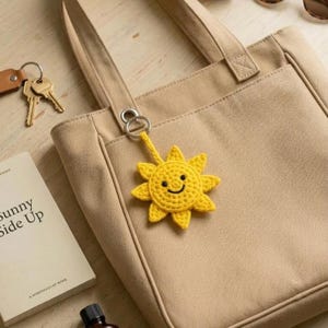 Puede incluir: Un bolso tote de lona beige con un llavero de sol de ganchillo amarillo adjunto. El sol tiene una cara sonriente. También se ve un libro titulado "Sunny Side Up" y un juego de llaves.