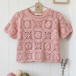 以下が含まれることがあります： 正方形のパターンデザインの、薄いピンク色の半袖かぎ針編みトップス。白い壁を背景に、木製のハンガーに吊るされています。「HANDMADE COLLECTION」と「AURA KNITS」と書かれたタグが付いています。