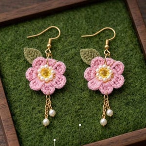 Può includere: Un paio di orecchini a fiori all'uncinetto fatti a mano. Ogni orecchino presenta un fiore rosa con un centro giallo e una catena dorata pendente con perle bianche. Gli orecchini hanno ganci per le orecchie color oro e accenti di foglie verdi.