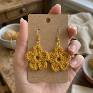 May include: A pair of handmade, mustard yellow crochet flower earrings with gold-colored hooks. The earrings are displayed on a brown card, held by a person. The background includes a wooden surface and a bowl with yarn.