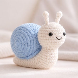 以下が含まれることがあります： 手作りの編み物カタツムリのおもちゃ。カタツムリは、水色の殻とクリーム色の体を持っています。黒い目と笑顔の口が特徴です。触角もクリーム色です。柔らかい糸で作られたおもちゃです。