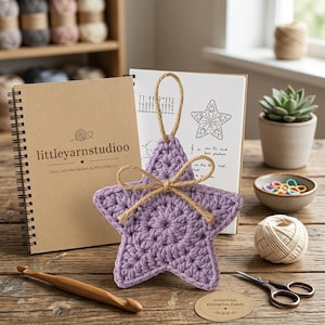 May include: A lavender crochet star ornament with a jute twine hanging loop and a tied bow. The star is positioned in front of a spiral-bound notebook labeled littleyarnstudioo, accompanied by a wooden crochet hook and metal scissors.