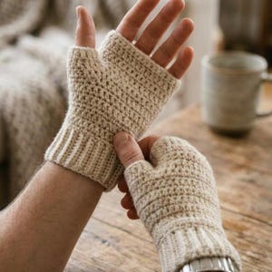 May include: Beige crochet fingerless gloves with ribbed cuffs. The gloves are made of a textured yarn and are designed to keep hands warm while leaving fingers free. A mug is in the background.