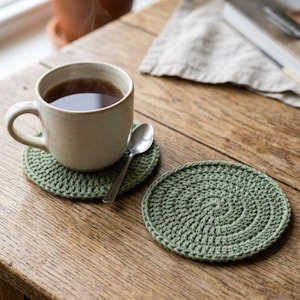 May include: A beige ceramic mug filled with tea sits on a green crocheted coaster, with a silver spoon resting on the coaster. Another green crocheted coaster is next to it. The items are on a wooden table.