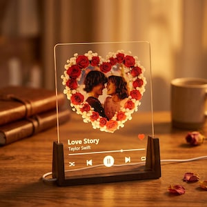 Op de afbeelding: Een rechthoekige, doorzichtige acrylplaat in een donkere houten voet, met een hartvormige bloemenkrans rond een foto van twee personen. Daaronder staat de tekst Love Story van Taylor Swift met iconen van een muziekspeler.