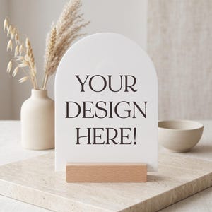 May include: A white arched sign with a smooth surface featuring the black text "YOUR DESIGN HERE!" printed in a serif font. The sign is held upright by a rectangular light-coloured wooden base stand, creating a clean and simple display piece.