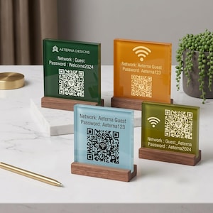 Könnte beinhalten: Vier quadratische WLAN-Schilder aus Acryl in Grün, Orange, Hellblau und Oliv, jeweils in einem rechteckigen Holzsockel. Die Schilder zeigen Netzwerknamen, Passwörter und QR-Codes. Ein goldener Stift liegt in der Nähe der Schilder.