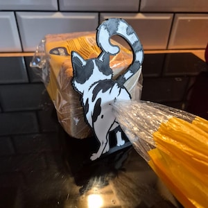 Puede incluir: Un clip para bolsas de pan con forma de gato, impreso en 3D en blanco y negro. El accesorio muestra a un gato estilizado visto desde atrás con una cola curva, diseñado para mantener cerrada una bolsa de plástico. La figura es plana.