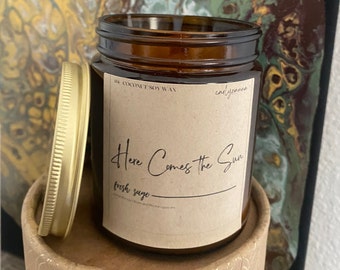 Puede incluir: Un tarro de vidrio marrón para velas con tapa dorada. La etiqueta dice "Here Comes the Sun" y "fresh sage". El tarro está sobre una caja de cartón marrón.