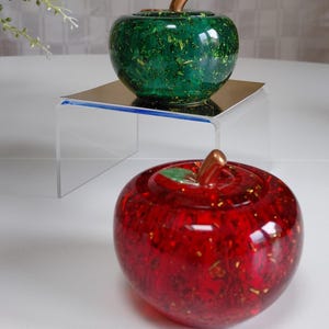 May include: Two decorative apple-shaped objects. One is green with gold flecks, the other is red with gold flecks. Both have bronze-coloured stems and are on a clear acrylic stand. The apples are on a white surface.