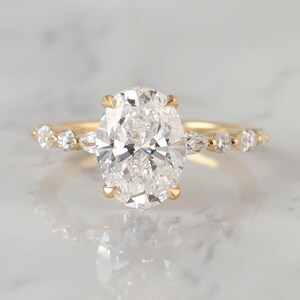 Puede incluir: Un anillo de oro amarillo con un gran diamante central de talla oval sostenido por cuatro garras. La banda está adornada con tres diamantes pequeños de talla redonda y un diamante de talla marquesa a cada lado, creando un diseño simétrico y detallado.