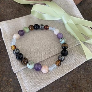 Peut inclure: Un bracelet composé de perles sphériques polies dans des tons de violet, noir, marron, bleu clair, vert et rose pâle. Le bracelet est disposé en un motif circulaire avec des perles de tailles et de couleurs alternées.