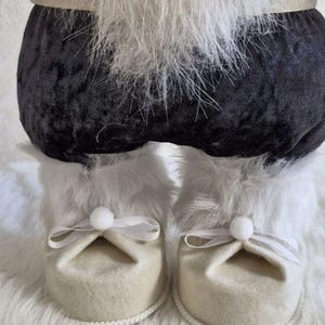 May include: A close-up of a decorative figure's lower body. The figure has black velvet pants, white faux fur trim, and cream-colored felt shoes with white bows. The figure is on a white faux fur surface.