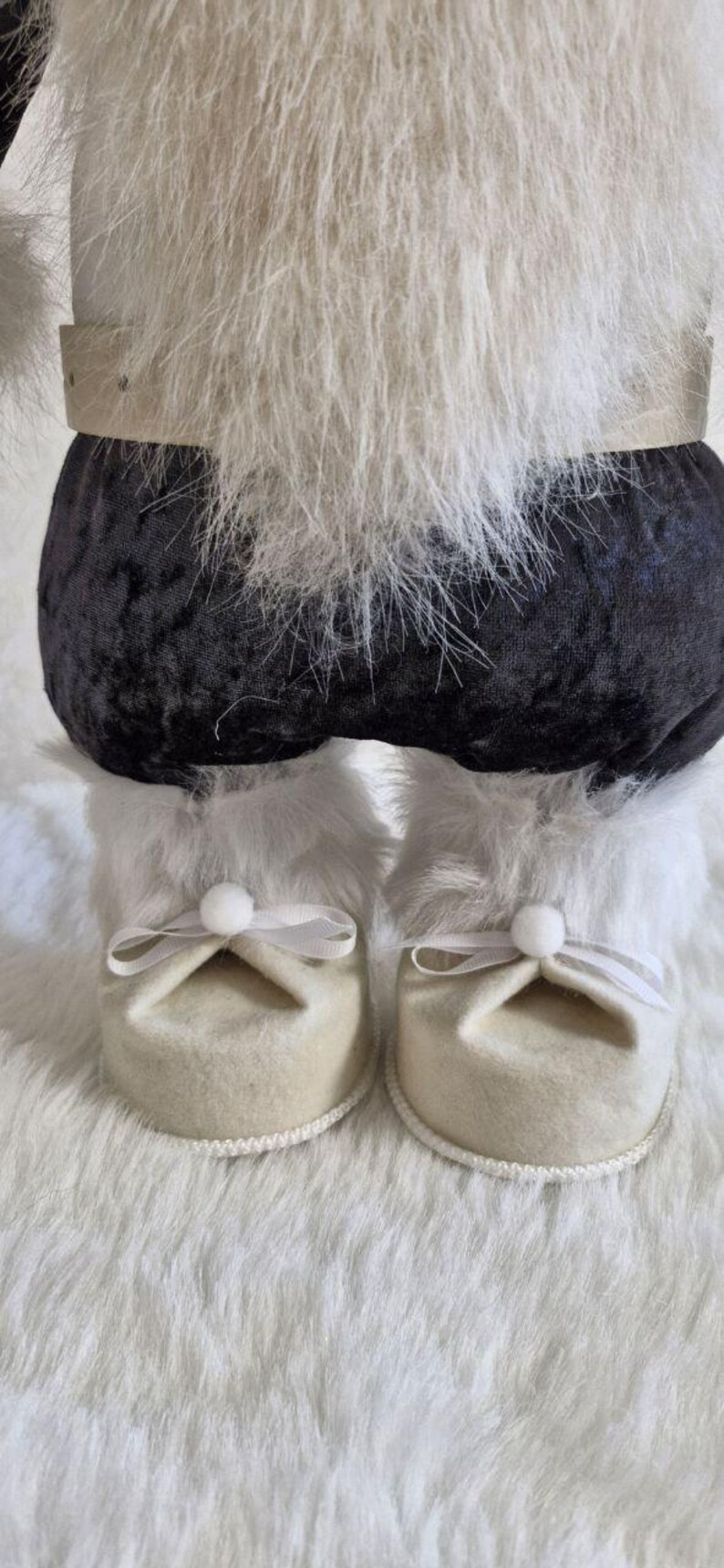 May include: A close-up of a decorative figure's lower body. The figure has black velvet pants, white faux fur trim, and cream-colored felt shoes with white bows. The figure is on a white faux fur surface.
