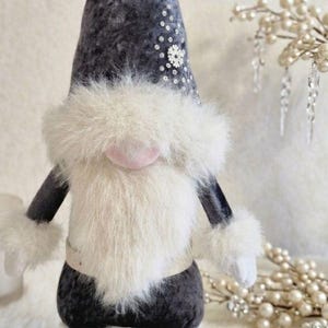 May include: A plush gnome doll with a dark gray body, arms, and hat. The hat is decorated with sequins and a snowflake. It has a large white fluffy beard, pink nose, and white fur cuffs. A decorative item for the holiday season.