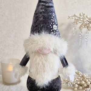 May include: A plush gnome doll with a dark gray velvet hat and body, adorned with white faux fur trim and a fluffy white beard. The hat is embellished with sequins. The gnome wears white shoes with bows and has a pink nose.