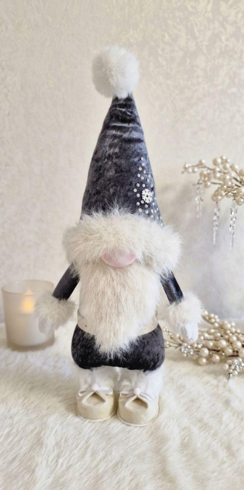 May include: A plush gnome doll with a dark gray velvet hat and body, adorned with white faux fur trim and a fluffy white beard. The hat is embellished with sequins. The gnome wears white shoes with bows and has a pink nose.