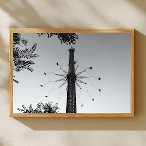 May include: Black and white photograph of a framed amusement park ride. The ride is a tall tower with swings extending outward. The sky is a light gray, and the frame is a light brown color.