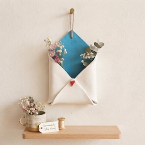 Puede incluir: Un bolsillo de pared de cerámica blanca con forma de sobre y un interior azul, con un pequeño corazón rojo. Cuelga de un lazo de yute y contiene flores secas. Una taza de cerámica con flores secas se encuentra sobre un estante de madera.