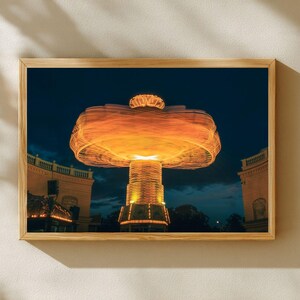 May include: A framed print of a carnival ride at night. The ride is illuminated with orange and yellow lights, creating a blurred effect from motion. The background is a dark blue sky with buildings on either side.